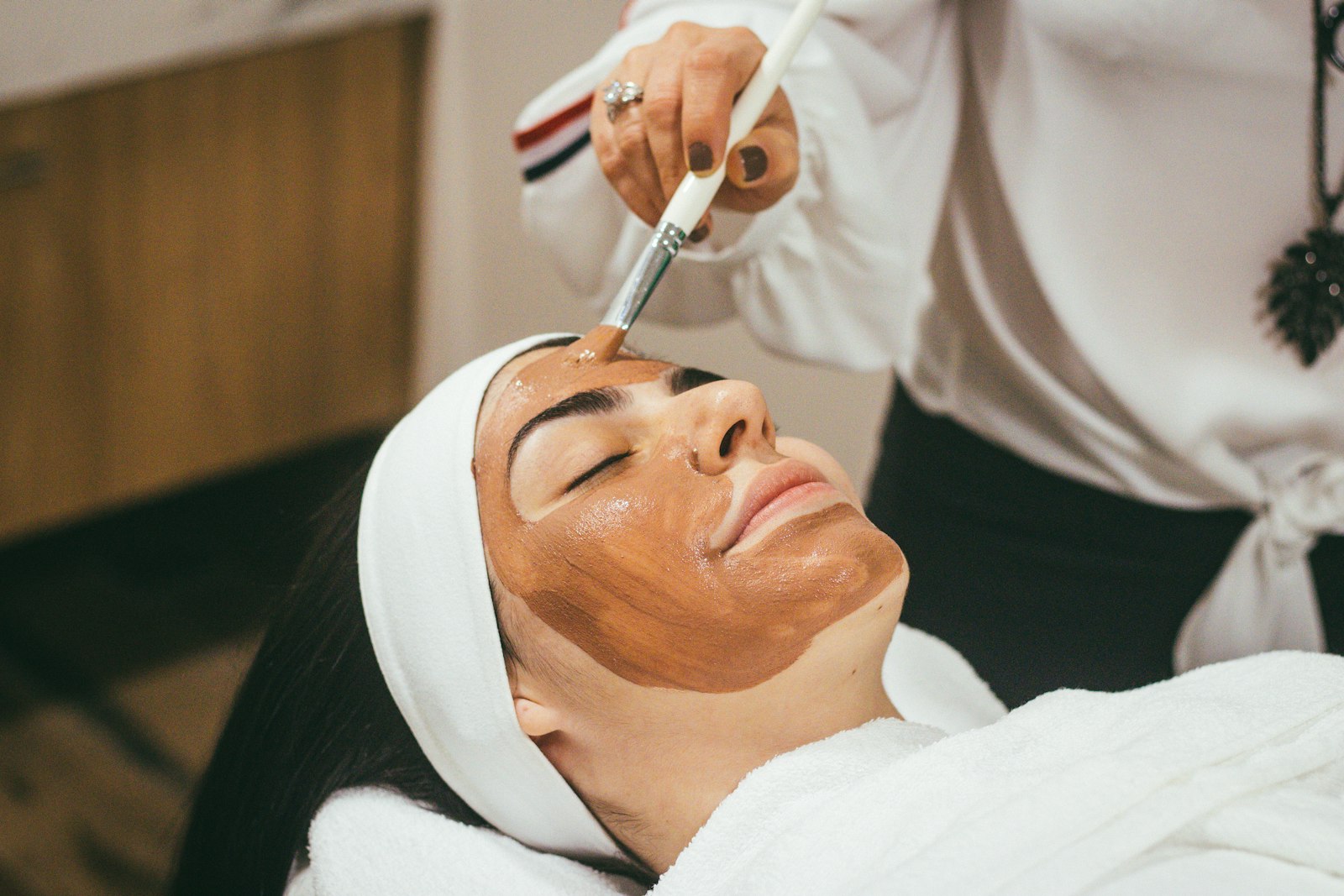 A beauty therapist applying a facial mask with a brush to a client wearing a headband