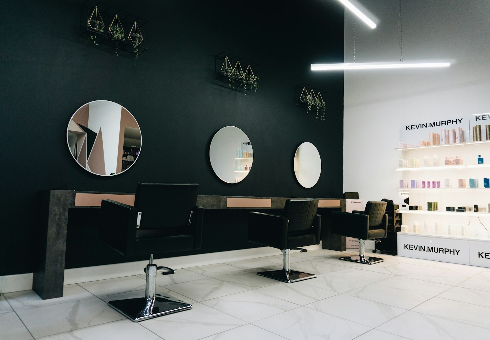 Three empty salon chairs facing round mirrors on a dark wall